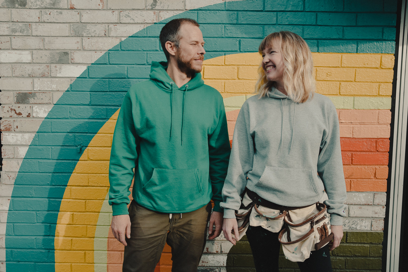 Two people wearing green and gray hoodies standing in front of a colorful wall.